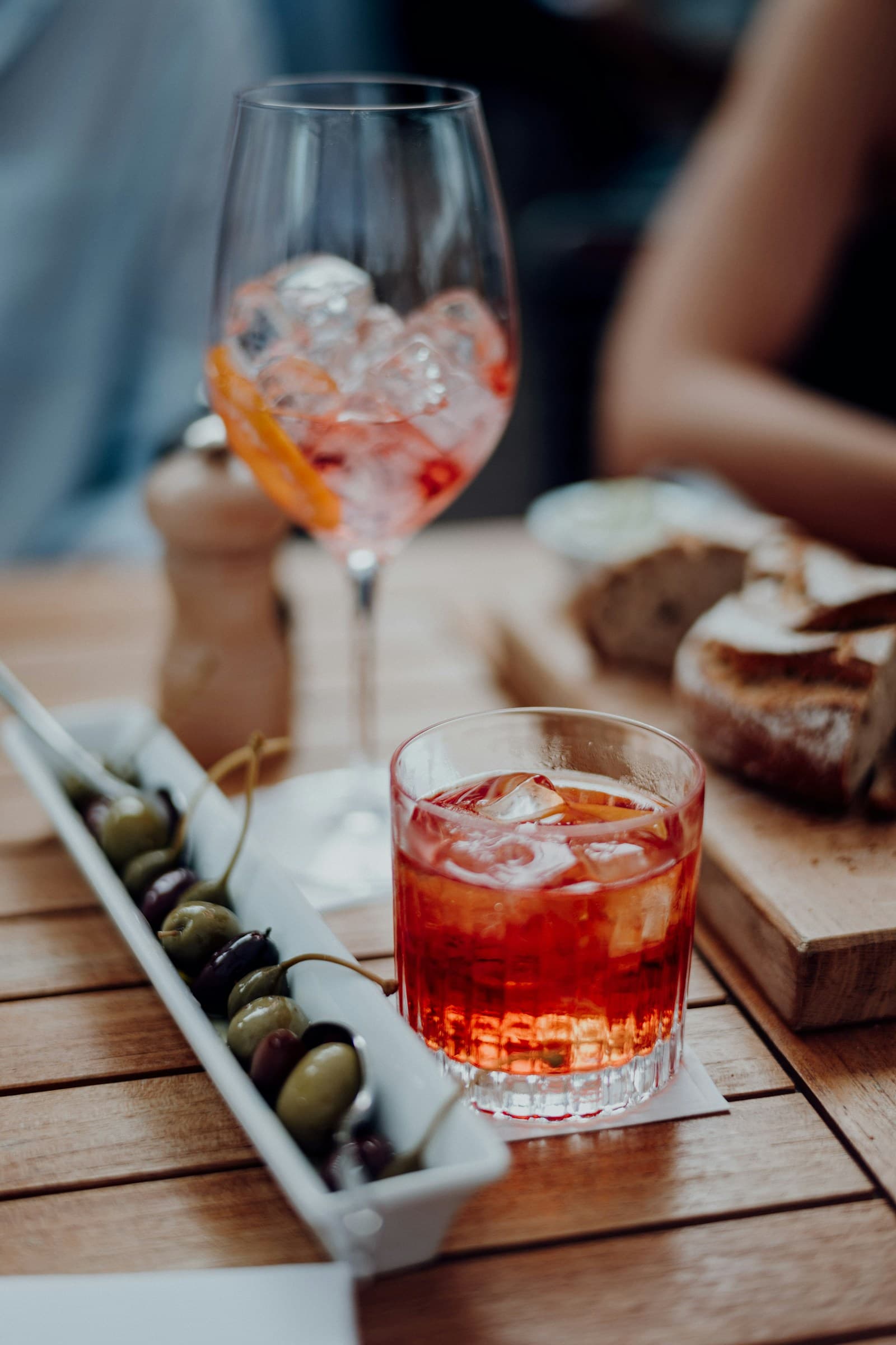 A negroni and an Aperol spritz beside a dish of olives and bread on a sunlit wooden terrace table