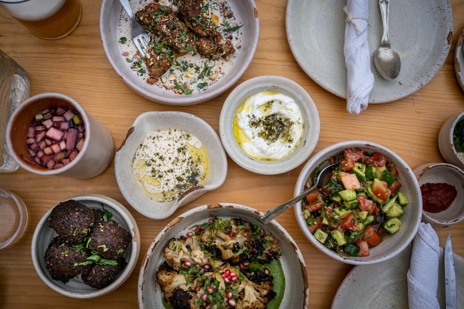 An overhead spread of Mediterranean plates to share on a warm wood table: falafel, dips, grilled cauliflower, a tomato and cucumber salad, olives
